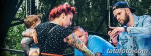 Фото тату: женщина с ребенком на руках и мужчина фотографируют татуировку на руке другого мужчины на тату-фестивале.
