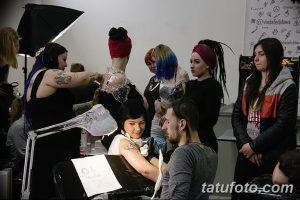 Фото тату: мастер тату делает татуировку девушке, вокруг наблюдают другие люди на тату-конвенции.