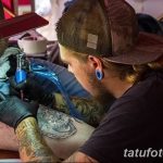 Фотография мастера тату, работающего над татуировкой на теле клиента. Видна тату-машинка, рука мастера с татуировками и часть тела клиента.