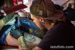 Фотография мастера тату, работающего над татуировкой на теле клиента. Видна тату-машинка, рука мастера с татуировками и часть тела клиента.