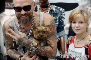 Фотография мужчины с обширными татуировками на руках и теле, держащего на руках маленького йоркширского терьера.