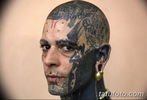 Фото мужчины с экстремальными татуировками на лице, голове и шее. У него пирсинг и необычный взгляд.