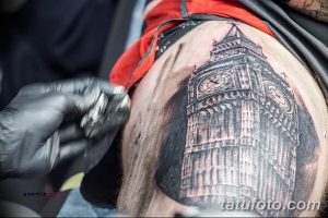 Фото тату процесса нанесения реалистичной черно-белой татуировки Биг-Бена на плечо. Мастер сосредоточенно работает над деталями циферблата.