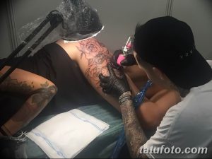 Фото тату процесса нанесения черно-белой татуировки розы на плечо. Мастер в кепке и черной майке работает над деталями лепестков.