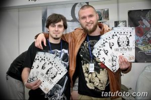 Фото двух мастеров тату на фестивале. Они держат в руках эскизы татуировок и улыбаются.