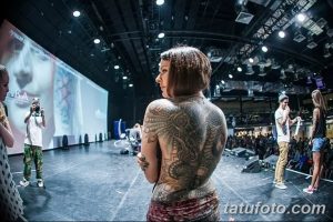 Фото тату на спине девушки на фестивале татуировок. Мастер демонстрирует работу, на заднем плане сцена и зрители.