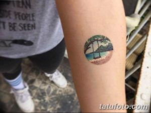 Фото тату на предплечье в форме круга с изображением девушки, сидящей на качелях под деревом на фоне пляжа.