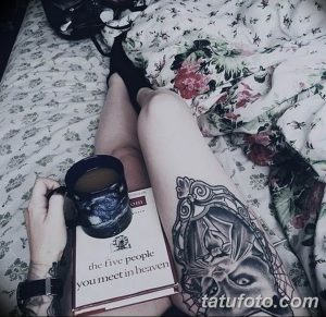 Фото тату на ноге девушки, читающей книгу. Рисунок татуировки изображает лицо ангела в стиле реализм с элементами готики и цветочным орнаментом.