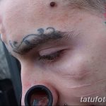 Фото татуировки с изображением усов над бровью и небольших цветов на виске. Оригинальный и экстравагантный рисунок.