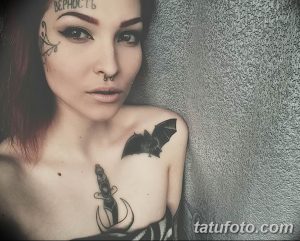 Фото девушки с татуировками на лице, шее и груди. На лбу видна надпись 'ВЕРНОСТЬ', на плече – изображение летучей мыши.