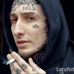 Фото татуировки на лице мужчины в капюшоне. Множество мелких татуировок, включая звезды, надписи и узоры, покрывают лоб, щеки и подбородок.