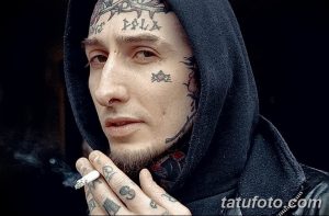 Фото татуировки на лице мужчины в капюшоне. Множество мелких татуировок, включая звезды, надписи и узоры, покрывают лоб, щеки и подбородок.