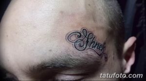 Фото тату: небольшая надпись 'Shnek' на лбу мужчины. Минималистичная татуировка черным цветом.