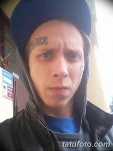 Фото татуировки на лице мужчины: надпись и узор расположены на лбу. Тату выполнено в черном цвете.