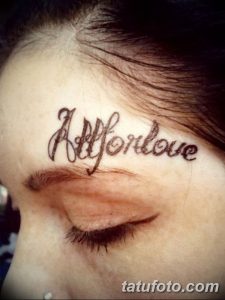 Фото тату над глазом девушки с надписью "All for love", выполненной каллиграфическим шрифтом. Изображение демонстрирует татуировку вблизи.