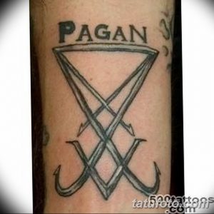 Фотография простой татуировки на руке с надписью 'PAGAN' над геометрическим символом, напоминающим перевернутый треугольник с крестом.