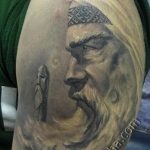 Фото тату: реалистичный портрет воина с длинными волосами и бородой на плече. Серые оттенки и детализированная проработка лица.