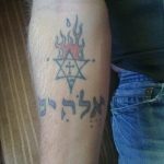 Фото тату на предплечье, изображающее Звезду Давида с огненным нимбом и надписью на иврите. Рисунок выполнен в черно-белых тонах.