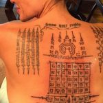 Фото тату: сложный геометрический рисунок с тайскими письменами и символами на спине мужчины. Традиционный стиль сакральной татуировки.