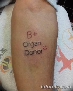 Фото тату на предплечье: надпись 'B+ Organ Donor' с изображением смайлика и сердечка. Татуировка выполнена в черном цвете.