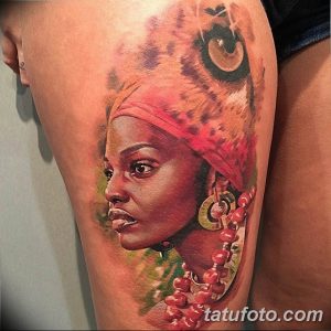 Фото тату: реалистичный портрет африканской женщины с головным убором и цветами на плече, выполненный в ярких цветах.