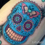 Фото тату сахарного черепа с бабочкой на плече. Яркий рисунок в стиле Day of the Dead с использованием голубых, розовых и оранжевых цветов.