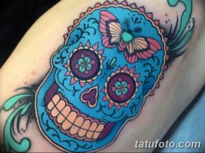 Фото тату сахарного черепа с бабочкой на плече. Яркий рисунок в стиле Day of the Dead с использованием голубых, розовых и оранжевых цветов.