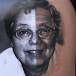 Фото тату с изображением портрета пожилой женщины в очках на руке. Черно-белый стиль, реалистичная проработка лица и деталей. Трогательный рисунок татуировки.