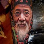 Фотография татуировки с реалистичным портретом пожилого мужчины с длинной бородой и красными маками, выполненной на руке.