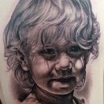 Фото тату детского портрета на руке. Черно-серый рисунок с детальной проработкой лица и нежной улыбки ребенка.
