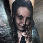 Фото тату женского портрета на руке. Черно-серый рисунок с детальной проработкой лица и волос, украшенных цветами.