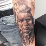 Фотография татуировки на руке с изображением портрета пожилого индейца в головном уборе, выполненного в реалистичном стиле с высокой детализацией.