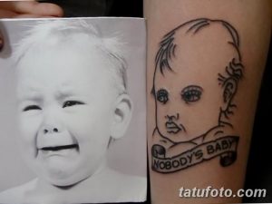 Фото тату детского портрета на руке. Рисунок выполнен в черно-белых тонах с добавлением декоративных элементов и надписи 'Nobody's Baby'.