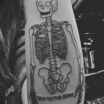 Фото тату: простой рисунок скелета на руке с надписью "BAD TO THE BONE", выполненный в стиле олдскул.