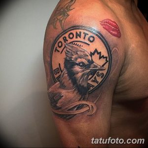 Фото татуировки на плече с изображением логотипа бейсбольной команды Toronto Blue Jays. Рисунок выполнен в черно-серых тонах с высокой детализацией.