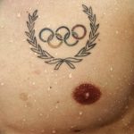 Фото тату минималистичного изображения олимпийских колец, обрамленных лавровым венком, на женской груди.