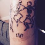 Фото татуировки двух танцующих скелетов с надписью "YAY!". Черно-белый рисунок татуировки с юмористическим подтекстом.