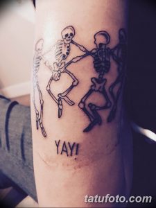 Фото татуировки двух танцующих скелетов с надписью "YAY!". Черно-белый рисунок татуировки с юмористическим подтекстом.