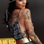 Фотография известной личности с множеством татуировок на спине, руках и шее, одетой в платье с геометрическим принтом.