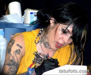 Фотография Kat Von D во время работы над татуировкой, с татуировками на руках и шее, в тату-салоне.