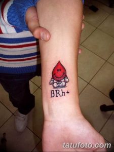 Фото тату: яркая капля крови с улыбающимся лицом и надписью 'BRh+'. Необычный и игривый рисунок татуировки на предплечье.