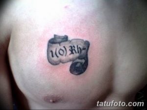 Фото татуировки на груди, изображающей свиток с надписью '1(0) Rh-', выполненной в серо-черных тонах.