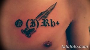 Фото тату на груди: патрон, обвитый веткой с листьями, и надпись O(II)Rh+, выполненное в черном цвете с детальной проработкой.
