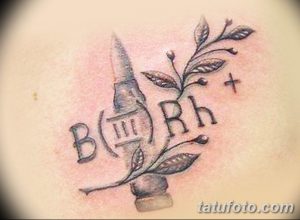 Фото тату на коже: патрон, обвитый веткой с листьями, и надпись B Rh+, выполненное в черном цвете с детальной проработкой.
