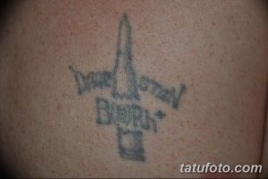 Фото старой, выцветшей татуировки в виде креста с патроном на спине. Черно-серый рисунок с размытыми контурами.