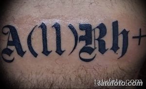 Фото тату: черная готическая надпись 'A(II)Rh+' на плече. Детализированный рисунок татуировки выполнен четкими линиями.