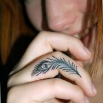 Фото татуировки пера на пальце. Минималистичный рисунок, выполненный черными чернилами, с детальной проработкой текстуры пера.