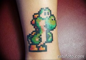 Фото тату в стиле 8-bit: персонаж Йоши из Super Mario Bros. на предплечье. Яркие цвета и пиксельная графика.