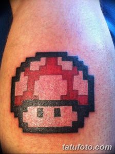 Фото тату пиксельного гриба из игры Super Mario на плече. Яркий рисунок в стиле 8-bit, выполненный в красных и черных тонах.