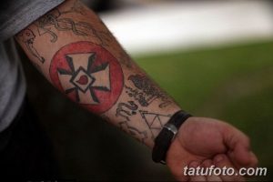 Фото тату на руке в стиле трайбл с изображением креста в круге и различных символов, выполненное в черном цвете с использованием геометрических узоров.